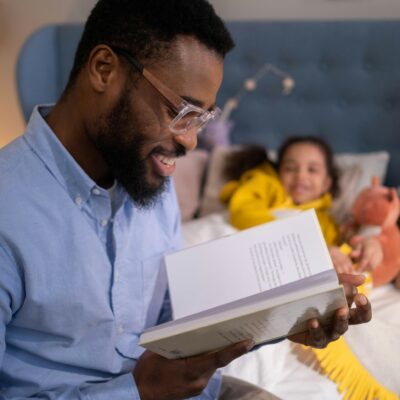 A man reads a book to his daughter, who is tucked in bed surrounded by stuffed animals