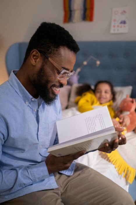 A man reads a book to his daughter, who is tucked in bed surrounded by stuffed animals